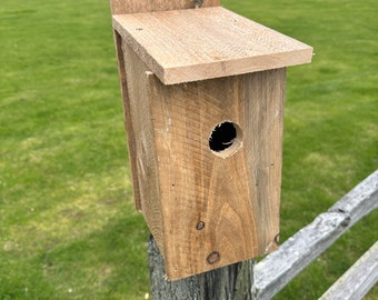 Puede incluir: Una casita de pájaros de madera con un pequeño agujero de entrada redondo, montada en un poste de madera desgastado. La casita de pájaros está pintada de color marrón claro y tiene un diseño sencillo.