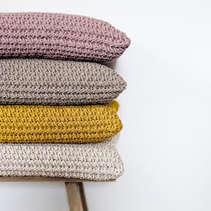 Pode incluir: Quatro almofadas de sofá de malha empilhadas em um banquinho de madeira. As almofadas são em tons de bege, marrom, amarelo e rosa.