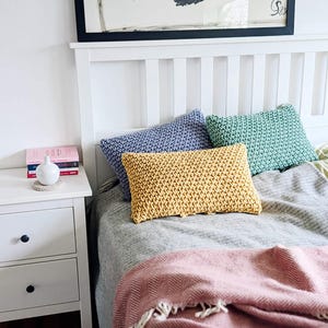 May include: A bed with a grey and white striped duvet cover and a pink throw blanket with fringe. There are three knitted throw pillows on the bed, one blue, one green, and one yellow.