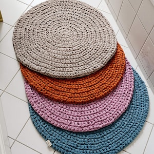 Könnte beinhalten: Vier runde gehäkelte Teppiche in verschiedenen Farben: Beige, Orange, Rosa und Blau. Die Teppiche sind übereinander gestapelt, wobei der beige Teppich oben liegt. Die Teppiche sind aus einem dicken, strukturierten Garn gefertigt.