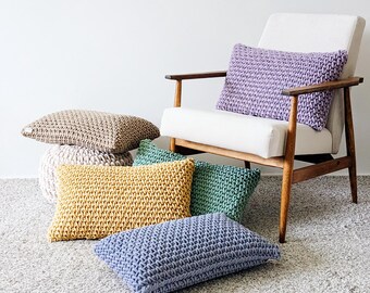 Könnte beinhalten: Eine Sammlung rechteckiger Strickkissen in verschiedenen Farben wie Lavendel, Senfgelb, Salbeigrün, Blau und Beige. Die Kissen weisen ein grobes, strukturiertes Strickmuster auf und sind um einen weißen Sessel mit Holzarmlehnen herum angeordnet.