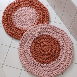 Könnte beinhalten: Zwei runde gehäkelte Badematten in Rosa und Terrakotta. Die Matten sind aus dickem Garn gefertigt und haben ein schlichtes, modernes Design.