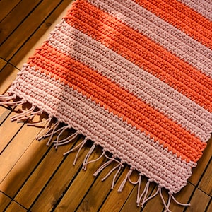 Peut inclure: Un tapis rectangulaire en crochet présentant des rayures horizontales alternées de fil orange vif et rose clair. Le textile possède un motif de mailles nouées texturé et est terminé par une frange en fil rose clair sur le bord inférieur.