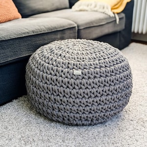 Könnte beinhalten: Ein grauer, gestrickter Pouf-Ottomane mit einem kleinen "indigo"-Etikett an der Seite. Der Pouf steht auf einem weißen, flauschigen Teppich vor einem grauen Sofa.