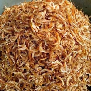 May include: A large pile of dried shrimp, with a mix of light and dark brown hues. The shrimp are small and curled, with visible legs and antennae. The image is a close-up, showing the texture and detail of the dried seafood.