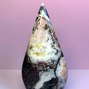 May include: A polished, teardrop-shaped stone sculpture featuring intricate, swirling patterns in shades of cream, white, dark brown, and black. The surface displays organic, concentric banding and mineral inclusions throughout the entire piece.