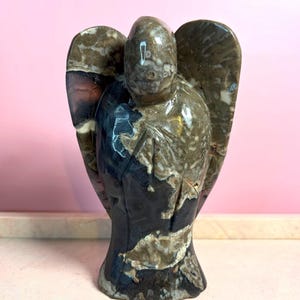 May include: A stone angel sculpture with outstretched wings. The statue features a mix of dark blue, brown, and cream colors. The angel has a smooth, polished finish and is set against a pink background. The sculpture is a decorative item.