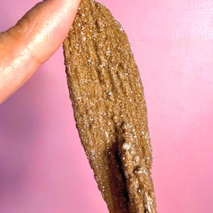 May include: A close-up of a brown mineral specimen with a textured surface and small, sparkling crystals. The mineral is held against a pink background, showcasing its natural color and unique formation. The mineral is approximately 4 inches long.