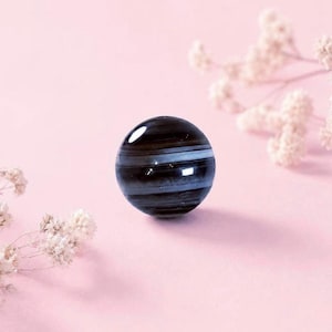May include: A spherical bead featuring a polished surface with horizontal bands of deep black and translucent white. The bead has a smooth, glossy finish that reflects light, creating bright highlights across its rounded, dark, and layered structure.