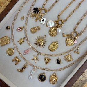 May include: Gold-toned necklaces with various charms, including a four-leaf clover, playing card, sun, heart, and seashell. The necklaces are displayed on a light gray surface.