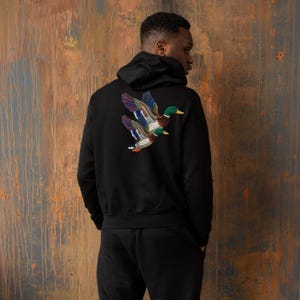 A man wearing a black hooded sweatshirt with a colorful embroidered duck design on the back.