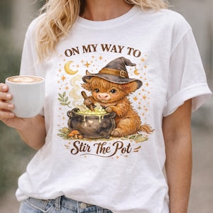 Puede incluir: Camiseta blanca con una vaca de las Tierras Altas de dibujos animados con un sombrero de bruja, removiendo un caldero. El texto "ON MY WAY TO" y "Stir The Pot" están impresos en la camiseta. El diseño incluye estrellas, una luna y follaje.