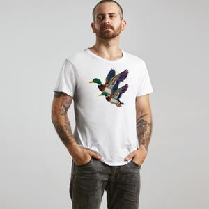 A man wearing a white t-shirt with a colorful embroidered duck design stands against a plain background.