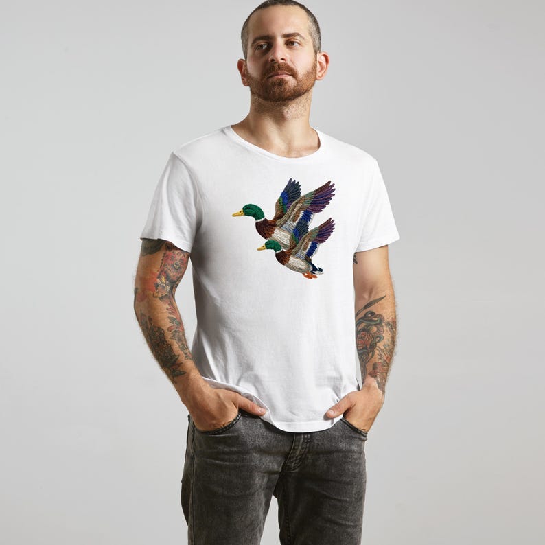 A man wearing a white t-shirt with a colorful embroidered duck design stands against a plain background.