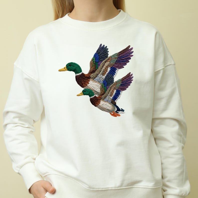 A white sweater with a colorful embroidered image of two ducks in flight.