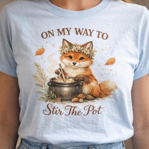 May include: Light blue t-shirt featuring a watercolor illustration of a fox wearing a flower crown, stirring a pot. The text "ON MY WAY TO Stir The Pot" is printed above and below the fox. The design includes leaves and mushrooms.
