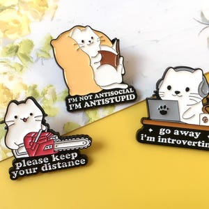 May include: Three enamel pins with white cat designs. One reads a book with the text "I'm not antisocial I'm antistupid." Another holds a chainsaw with "please keep your distance." The last uses a laptop with "go away I'm introverting."