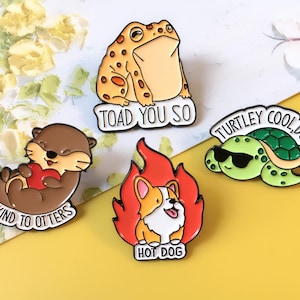 Puede incluir: Cuatro pines de esmalte con diseños de animales. Una nutria marrón sosteniendo un corazón rojo con el texto "BE KIND TO OTTERS". Una rana beige con el texto "TOAD YOU SO". Una tortuga verde con gafas de sol con el texto "TURTLEY COOL DUDE". Un corgi en llamas con el texto "HOT DOG".