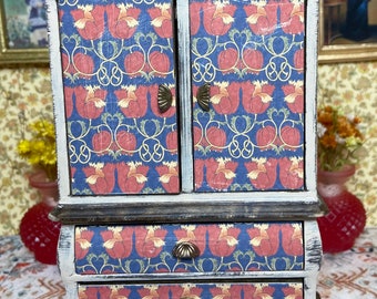 May include: A miniature wooden cabinet with two doors and two drawers. The cabinet is painted white with a distressed finish and has a dark brown trim. The doors and drawers are decorated with a blue floral pattern with red flowers and gold accents. The cabinet has brass-colored hardware.