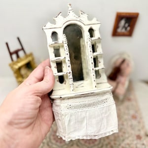 May include: A small, off-white decorative cabinet with a distressed finish. It features a mirrored back, small shelves, and a lace-trimmed skirt. The cabinet is held in a hand, showcasing its ornate design.