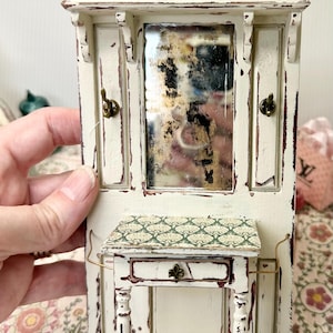 May include: A miniature, distressed white hall tree with a mirror, small table, and decorative hooks. The table has a patterned top and a drawer with a decorative pull. The piece has a vintage, shabby chic aesthetic.