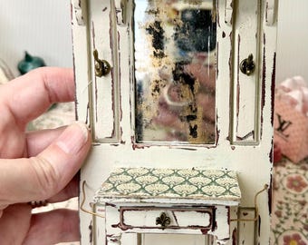 May include: A miniature, distressed white hall tree with a mirror, small table, and decorative hooks. The table has a patterned top and a drawer with a decorative pull. The piece has a vintage, shabby chic aesthetic.