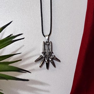 Puede incluir: Un collar con colgante plateado con un diseño geométrico cuelga sobre un fondo blanco. El collar tiene un cordón negro. Una planta verde en maceta y una tela burdeos añaden color a la escena.