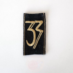 May include: A black rectangular patch with the gold number "33" embroidered on it. The edges of the patch are also trimmed with gold thread. The patch is set against a white background.