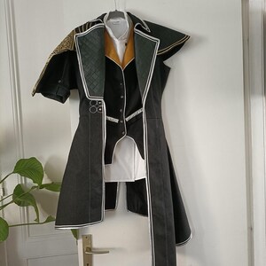 Peut inclure: Veste de costume noire et blanche avec des détails dorés, suspendue à un cintre. La veste présente un motif matelassé sur les épaules et les revers, avec une chemise blanche et un gilet doré en dessous. La veste a un design long et fluide.