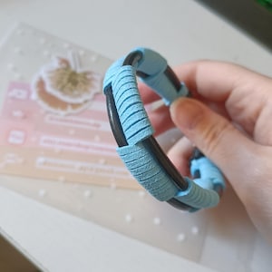 May include: A light blue and black bracelet with a braided design. The bracelet is made of leather and has a suede-like texture.