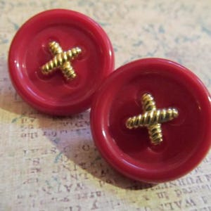 Può includere: Due orecchini a bottone rossi con un design a croce dorata al centro. Gli orecchini sono rotondi e hanno una finitura lucida. I bottoni sono un design classico, adatti a vari progetti di artigianato o come gioielli unici.