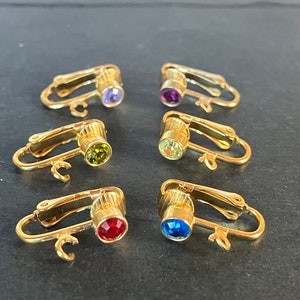 Op de afbeelding: Zes gouden clip-on oorbellen met verschillende gekleurde strass steentjes. De oorbellen zijn in een rij op een donker oppervlak gerangschikt. De strass steentjes zijn paars, groen, geel, rood en blauw.