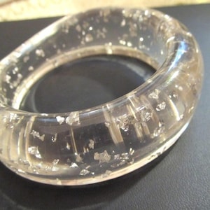 May include: A clear, thick resin bangle bracelet with an irregular, organic shape. The transparent material is embedded with numerous small, reflective silver-colored metallic flakes distributed throughout the entire body of the circular accessory.