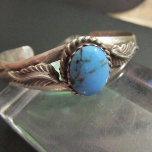 May include: A silver cuff bracelet featuring a central oval turquoise stone with dark matrix veining. The stone is set in a twisted rope border, flanked by embossed leaf motifs on the polished silver band, which has an open back design.