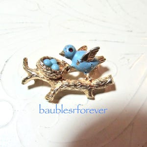 May include: A gold-tone metal brooch featuring a textured tree branch, a nest with three small blue eggs, and a light blue bird with a red eye. The text "baublesrforever" is printed in blue below the brooch on the white surface.