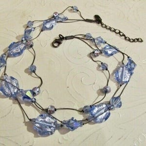 May include: A multi-strand necklace featuring light blue, faceted glass beads with a metallic finish. The necklace is constructed with thin, dark-toned metal chains and includes a lobster claw clasp and an adjustable extender chain for length.