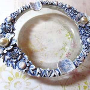 May include: A clear glass ashtray with a decorative blue and white floral border. The border features sculpted flowers and pearl accents. The ashtray has three built-in rests for cigarettes.