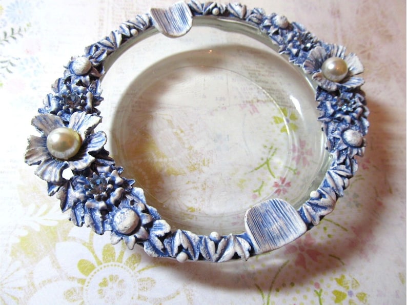 May include: A clear glass ashtray with a decorative blue and white floral border. The border features sculpted flowers and pearl accents. The ashtray has three built-in rests for cigarettes.