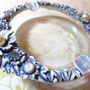May include: A decorative ashtray with a clear glass center and a floral border. The border is detailed with blue and white flowers, faux pearls, and small crystals. The ashtray has a vintage aesthetic.