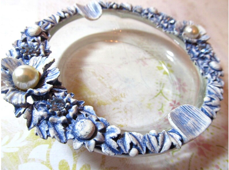 May include: A decorative ashtray with a clear glass center and a floral border. The border is detailed with blue and white flowers, faux pearls, and small crystals. The ashtray has a vintage aesthetic.
