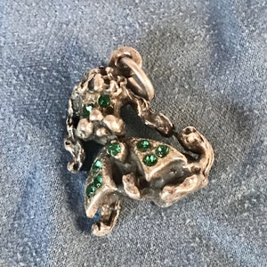 May include: A silver-toned pendant in the shape of a mythical creature, possibly a griffin or similar. The pendant is adorned with several small, round, emerald-green gemstones. The pendant has a loop at the top for attaching to a chain or bracelet.