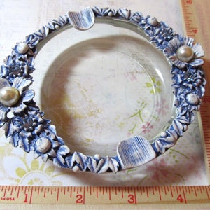 May include: A clear glass ashtray with a decorative blue and white floral border. The border features sculpted flowers and pearl-like accents. The ashtray has three built-in rests for cigarettes.