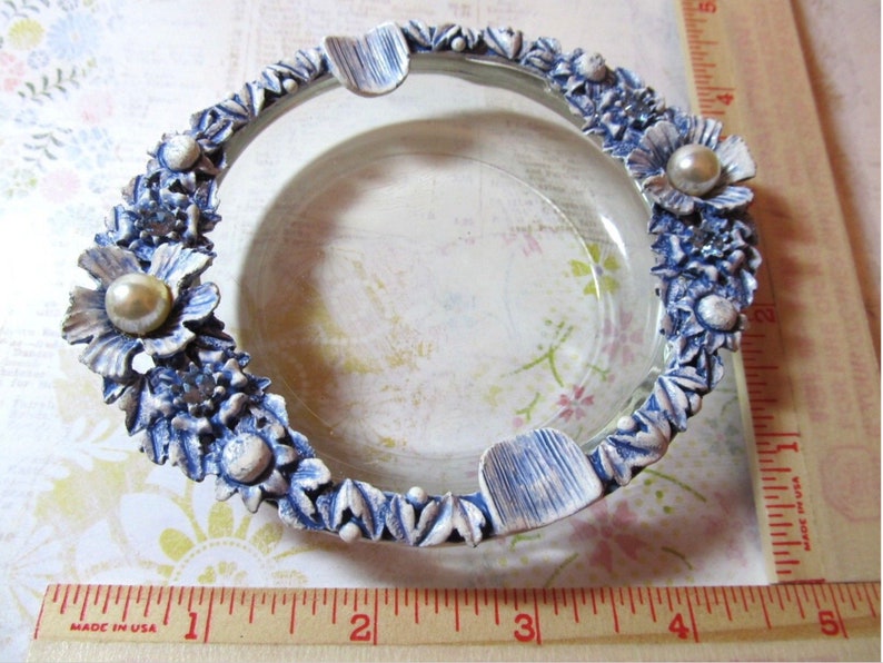 May include: A clear glass ashtray with a decorative blue and white floral border. The border features sculpted flowers and pearl-like accents. The ashtray has three built-in rests for cigarettes.