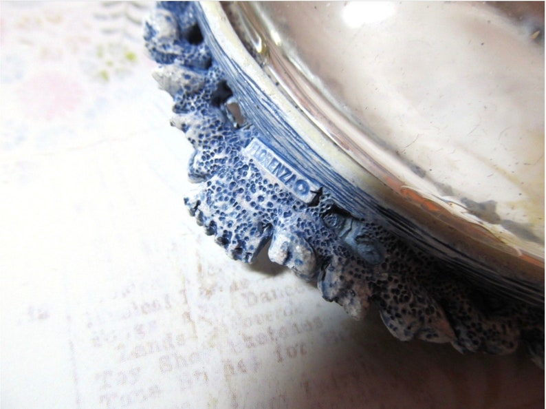 May include: Close-up of a decorative dish with a blue and white textured edge. The edge has a detailed, coral-like design. The word "FLORENZAO" is visible on a small rectangular label. The dish appears to be made of ceramic or a similar material.