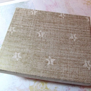 May include: A square box with a textured, gold-colored surface and a repeating pattern of white stars. The box is closed and appears to be made of cardboard or a similar material. The box is sitting on a light-colored surface.