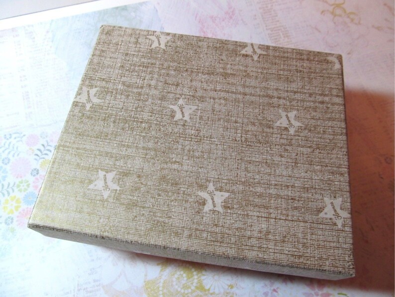 May include: A square box with a textured, gold-colored surface and a repeating pattern of white stars. The box is closed and appears to be made of cardboard or a similar material. The box is sitting on a light-colored surface.
