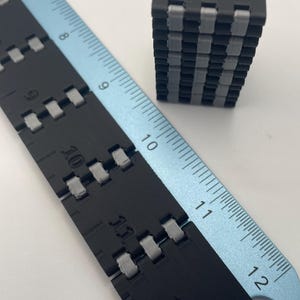 May include: A black and grey articulated ruler with measurements in inches, alongside a small, black and grey cube. The ruler is made of connected segments, and the cube is a stack of similar segments.