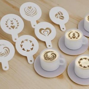 May include: A collection of white latte art stencils featuring heart, leaf, and coffee bean designs. These are placed near small white coffee cups, each with latte art, on light purple saucers. A coffee-themed arrangement.