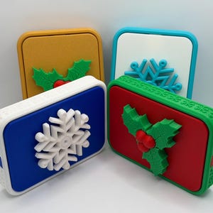 May include: Four rectangular power banks with festive designs. One has a yellow base with a green holly, another a blue base with a white snowflake, a third a red base with green holly, and the last a white base with a blue snowflake. Each has a different coloured border.