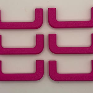 May include: A set of six bright magenta U-shaped drawer pulls arranged in two vertical columns of three. Each handle features a textured surface and a consistent, rounded rectangular profile, designed for attachment to furniture or cabinetry.